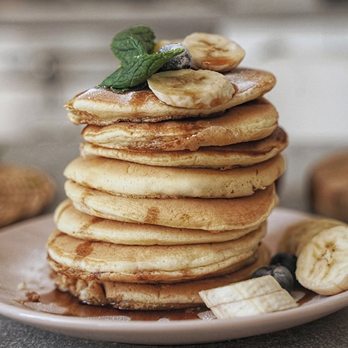 Photo of a stack of fluffy, maple-syrupy/caramely pancakes topped and plated with bananas and blueberries against an unfocused/blurred kitchen background - Olena Sergienko on Unsplash