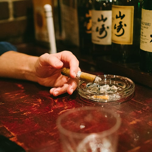 Photo of a man in a dark blue flannel (sleeve rolled up) holding a cigar over an ash tray with a row of Japanese malt whiskey bottles on a shelf - Julian Lozano on Unsplash