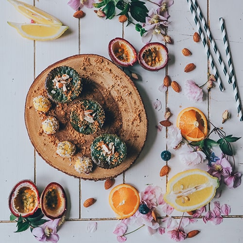 Photo of an assortment of foods, including almonds, hollowed-out dragonfruits (I think), citrus slices, and what looks like green açai bowls with coconut balls on a bark-like, wooden cutting board, surrounded by flowers and three green-and-white-striped paper straws  - Toa Heftiba on Unsplash