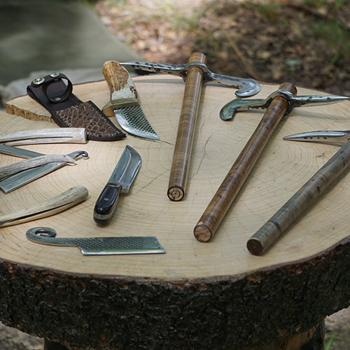Photo of a an assortment of weapons/tools (most notably knives and axes/picks) layed out on a slab of tree bark (repurposed as a table of sorts) - Chris Chow on Unsplash