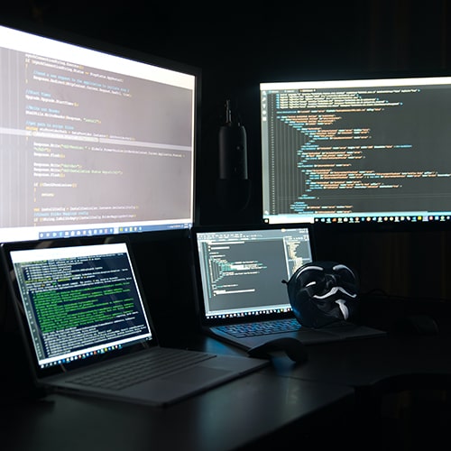 Image of computer monitors and laptops with coding programs open and lines of code on the screen, along with a black and white (inverted) 'Anonymous/Anon' mask - Clint Patterson on Unsplash