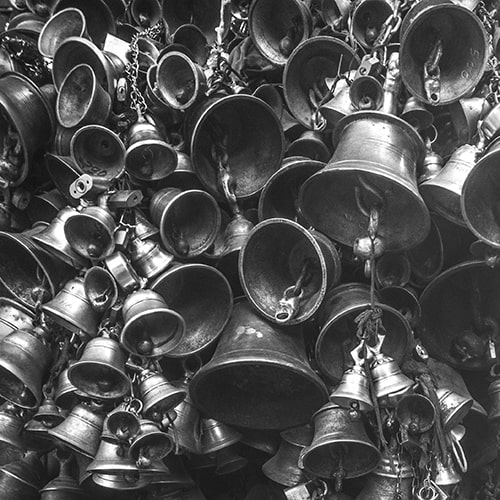 Black and white photo of a collection of bells of all sizes, hanging one on top of another in a giant collective - Shaouraav Shreshtha on Unsplash