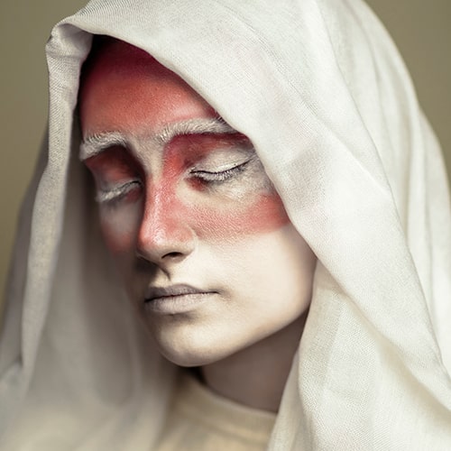 Photo of a woman with closed eyes in a sheer, white hooded cloak with red and white makeup on her face - Emad Kolahi on Unsplash