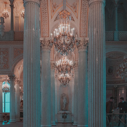 Photo of the interior of a castle, with columns and chandeliers in pink and teal lighting - Winter Castle interior - Francesco La Corte on Unsplash