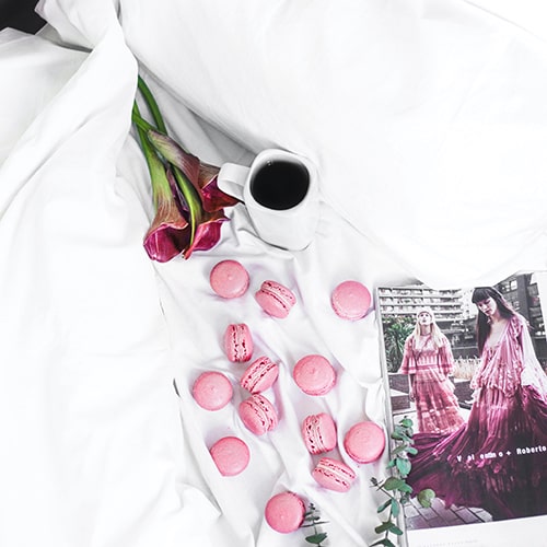 Photo of a cup of black coffee, pink tulip flowers, pink macaron,s and an open magazine on a white comforter - Alexandra Gorn on Unsplash