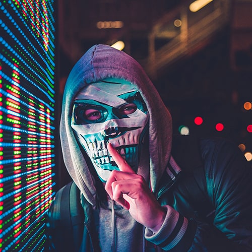Individual wearing an abstractly painted skull mask dressed in a hoodie and jacket leaning against a virtual display board while shushing the viewer.