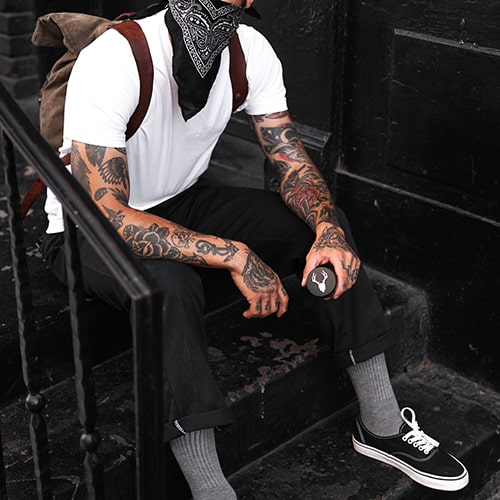 Preppy grunge male with sleeve tattoos and a bandana mask sitting on a distressed stoop.
