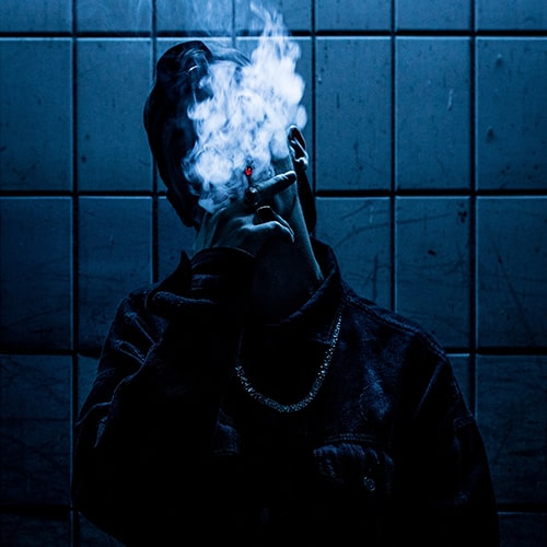 Moody blue shot of an anonymous, obscured male smoking and leaning against a tiled wall.