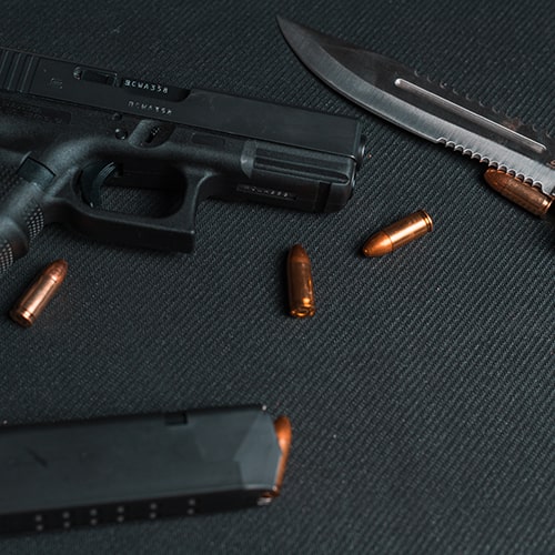 Weapons (black handgun and separate magazine with rust-colored bullets, and a hunting knife) placed on a black, textured piece of fabric.