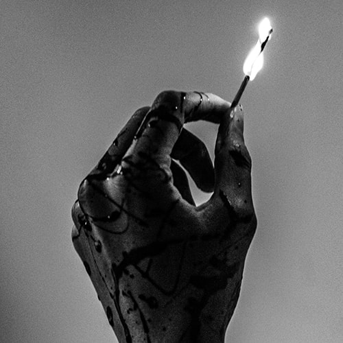 Black and white close-up of a hand, splatterd with some kind of dark liquid (possibly ink) holding a lit match.
