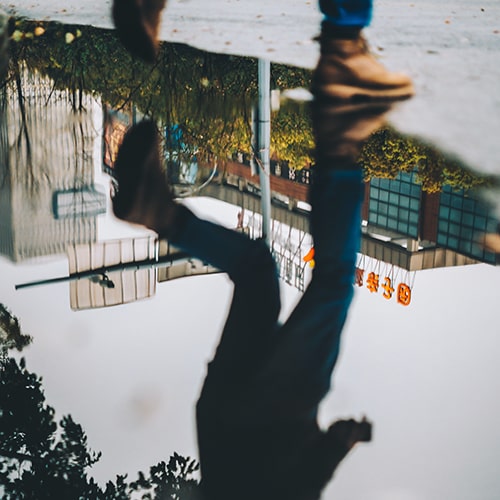 Male in brown leather boots walking by a puddle of water, with the main focus of the photo being on his reflection in the water.