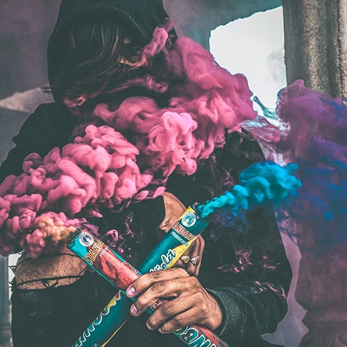 Anonymous male obscured by two lit colored smoke bombs (pink/magenta and blue).