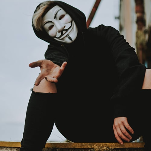 Blond guy with a Guy Fawkes Anonymous mask wearing all black, sitting down and holding his hand out invitingly.