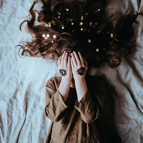 Photo of a brunette girl in a brown dress lying on a bed with star confetti in her hair, and she's covering her eyes - Annie Spratt on Unsplash