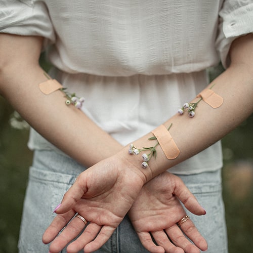 Photo of a girl with her arms crossed behind her back, hands crossed over one another, with bandaids on her arms with flowers underneath - Taisiia Stupak on Unsplash
