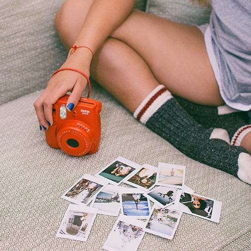 Photo of a girl sitting on a couch with a red-orange Instax Polaroid camera with a collection of developed polaroid pictures - Martin López on Unsplash