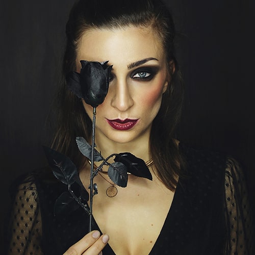 Photo of a woman with dark makeup and a sheer-sleeved black shirt holding a black rose up over her right eye - Apostolos Vamvouras on Unsplash