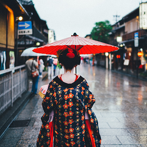 Photo of a Japanese woman dressed in a kimono as a geisha, back to the camera and with a red umbrella held over her head, walking down the street - Tianshu Liu on Unsplash