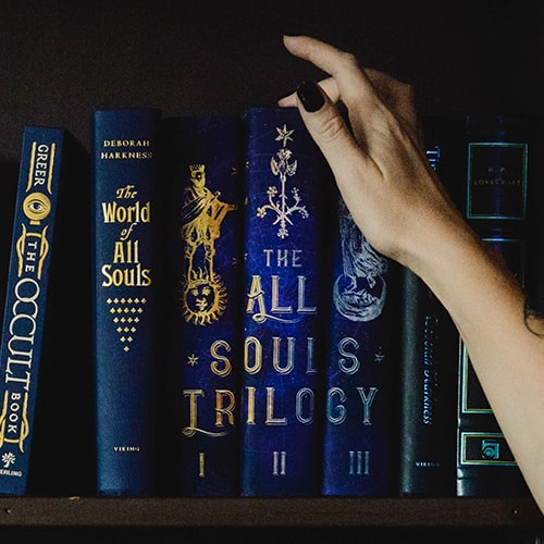 Photo of a woman's hand reaching to pick a book off a bookshelf, with witch/occult genre books across the shelf, some of which are titled 'The Occult Book', 'The World of All Souls', 'The All Souls Trilogy', etc. - Loren Cutler on Unsplash