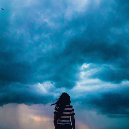 Photo of a girl standing in the foreground with storm clouds and rain in the background - Shashank Sahay on Unsplash