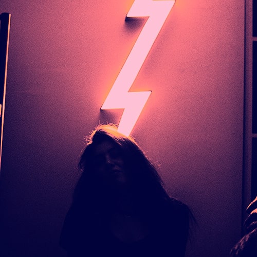 Dark, monochrome image of a woman sitting in front of a neon lightning bolt sign - Pars Sahin on Unsplash (edited by me)