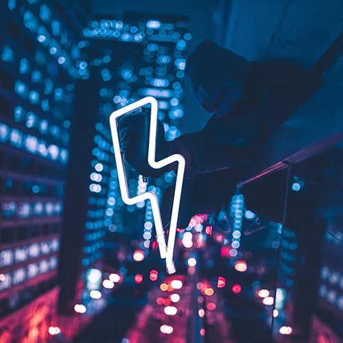Photo of a man in a blurred, bokeh-fied cityscape background holding a neon tube sign shaped like a lightning bolt - Max Bender on Usplash