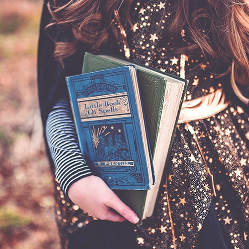 Girl in a foil star-decorated top and cape carrying two books, one of which has the title 'Little Book of Spells' - Paige Cody on Unsplash