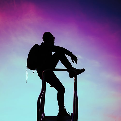 Silhouette of a man with a backpack and glasses on the top of a slide against a sunset sky