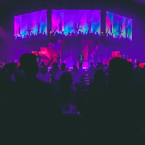 Photo of a band performing on a stage with three 'jumbotron' monitors livestreaming, photograph taken from the crowd - Outcast India on Unsplash