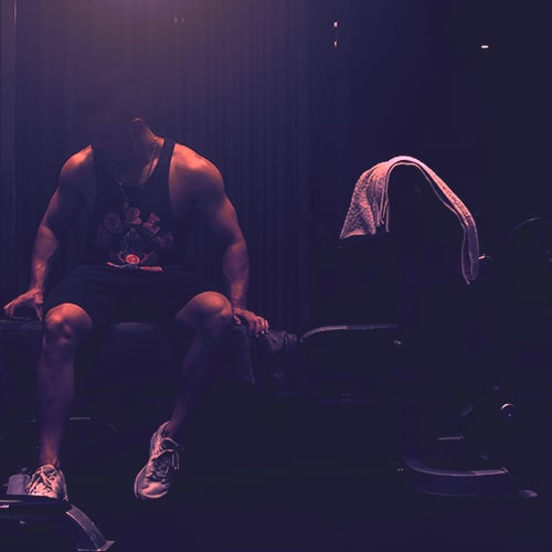 Photo of a man wearing a black tank top and shorts sitting on a weight bench with a tower over the headrest - Jaiden Peters on Unsplash