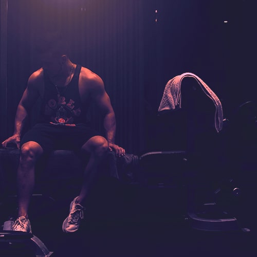 Photo of a man wearing a black tank top and shorts sitting on a weight bench with a tower over the headrest