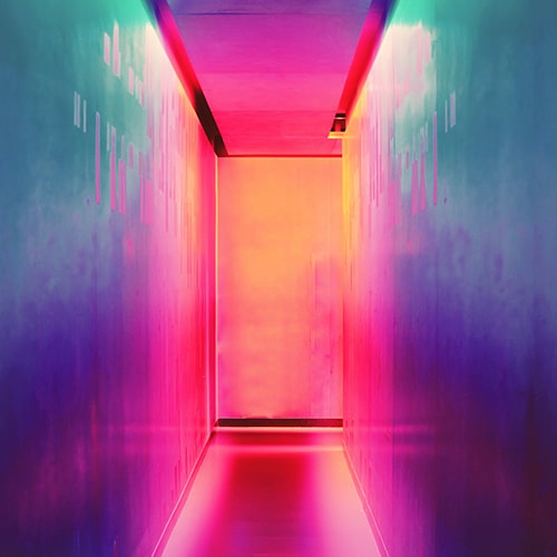Photo of an abstractly-lit hallway, with lights in oranges, reds, pinks, teals/blues, and purples