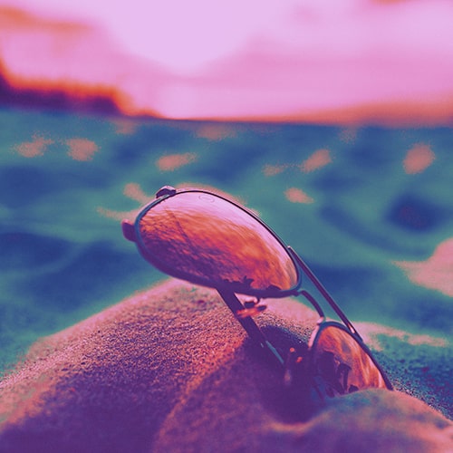 Close-up shot of a pair of aviators stuck in the sand against a blurred/unfocused backgrounf of water and a sunset - Alex Perez on Unsplash