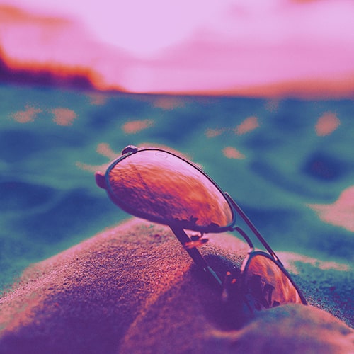 Close-up shot of a pair of aviators stuck in the sand against a blurred/unfocused backgrounf of water and a sunset