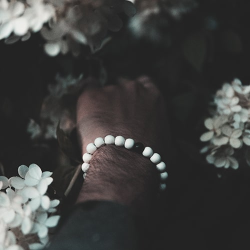 Photo of a man with a white beaded bracelet (with a sinle black bead in the middle), his hand amongst a bush of white flowers