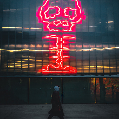 Photo of a man with a hooded coat walking in front of a reflective building, with a reflection in the windows of a neon light cartoon-like explosion with a skull-shaped smoke cloud