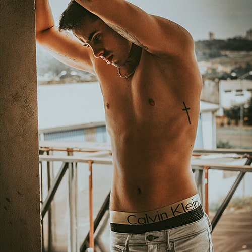 Photo of a shirtless man with a cross tattoo on his ribs posing with his arms over his head against a wall