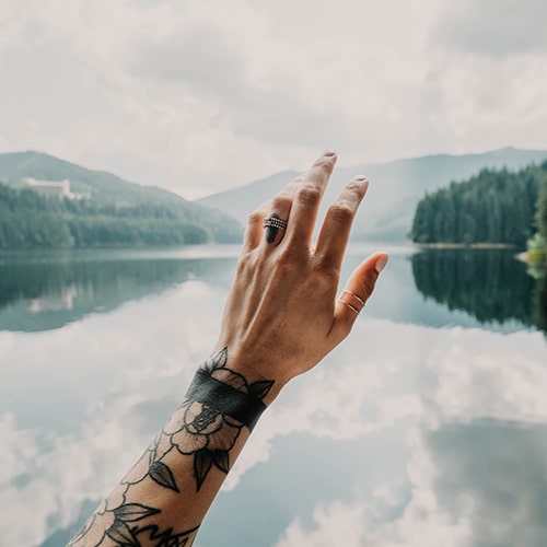 Photo of a man's sleeve-tattooed arm held up over an unfocused lake background