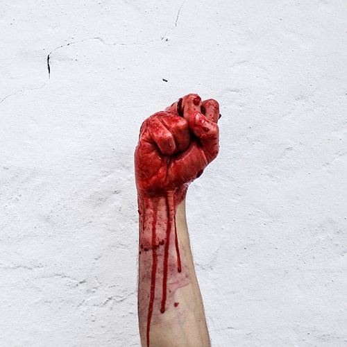 Photo of a fist raised against a white stone/concrete background, covered and dripping in a liquid/paint that's meant to resemble blood