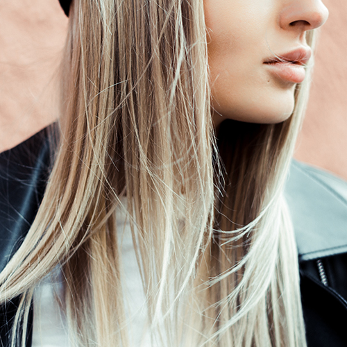 Photo of the lower half of a blonde woman's face who's wearing a white shirt and black leather jacket, against an blurry/unfocused pink textured wall background