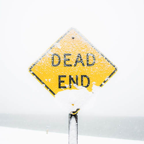 Photo of a snow-dusted yield sign that reads 'Dead End' against a blurred/unfocused background of snow and water