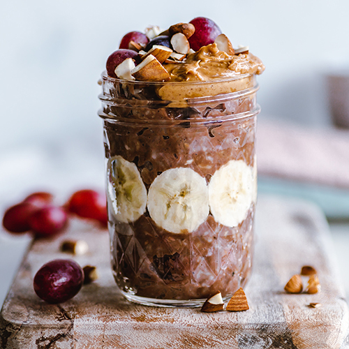Photo of a large, decorative glass jar filled with a chocolate-like, ganache-looking cream, bananas, walnuts, and fruit, on a wooden board