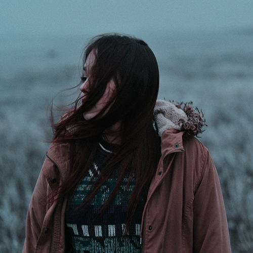 Photo of a woman in a winter jacket and sweater, wind blowing her hair in her face, against a blurred/unfocused wintry background - Iwan Shimko on Unsplash
